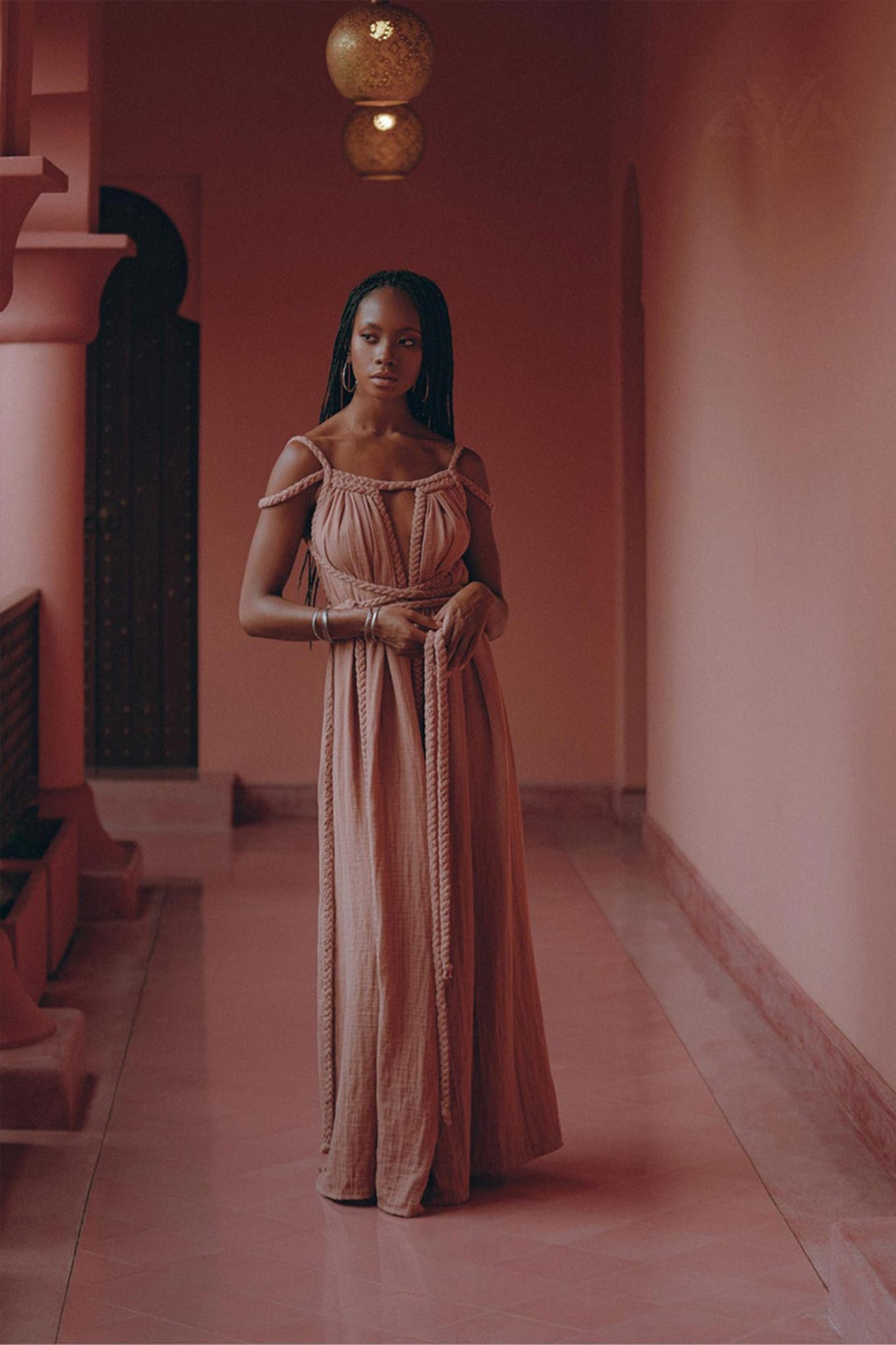 A woman wearing a Dusty Pink Greek Goddess Prom Boho Dress from AYA Sacred Wear stands in a dusty pink corridor adorned with ornate archways and hanging lanterns. The polished floor gleams beneath her, and a wooden bench sits to one side. With a serene expression, she gazes forward, embodying effortless elegance.