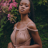 In front of a vibrant green background adorned with bright pink flowers, a woman with long braided hair confidently poses for the camera. She's wearing the Dusty Pink Greek Goddess Prom Boho Dress from AYA Sacred Wear, featuring stylish rope accents. Her look is enhanced by hoop earrings and natural makeup.