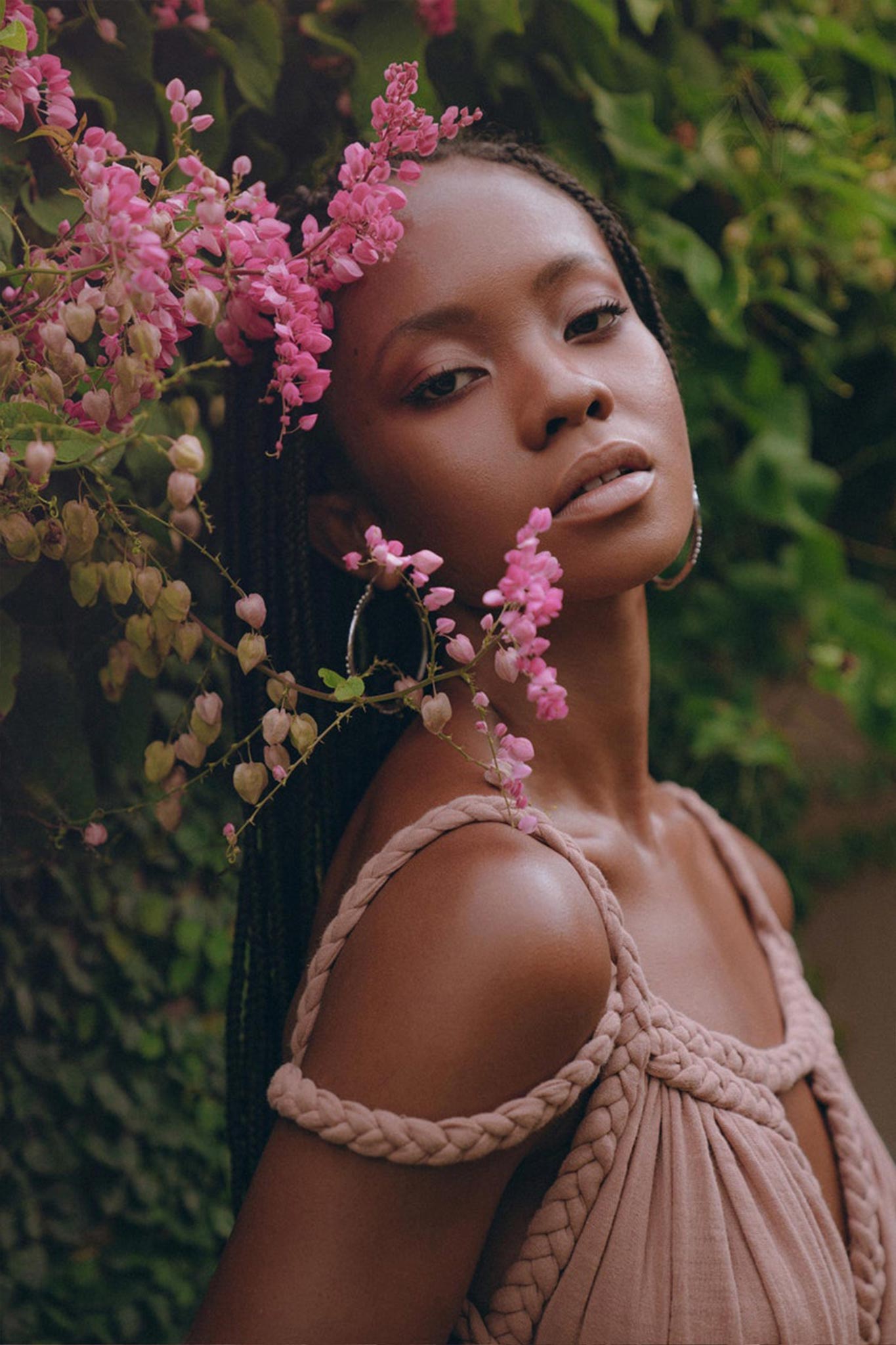 A woman with braided hair stands amidst pink flowers, wearing the Dusty Pink Greek Goddess Prom Boho Dress by AYA Sacred Wear, crafted from organic cotton. She gazes calmly at the camera, and the lush greenery in the background accentuates the serene beauty of the scene.