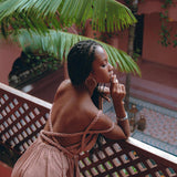 A woman with braided hair leans on a wooden railing, gazing thoughtfully into the distance. She is wearing a strappy brown dress that calls to mind the Dusty Pink Greek Goddess Prom Boho Dress by AYA Sacred Wear. The background features tropical plants and dusty pink architectural elements.