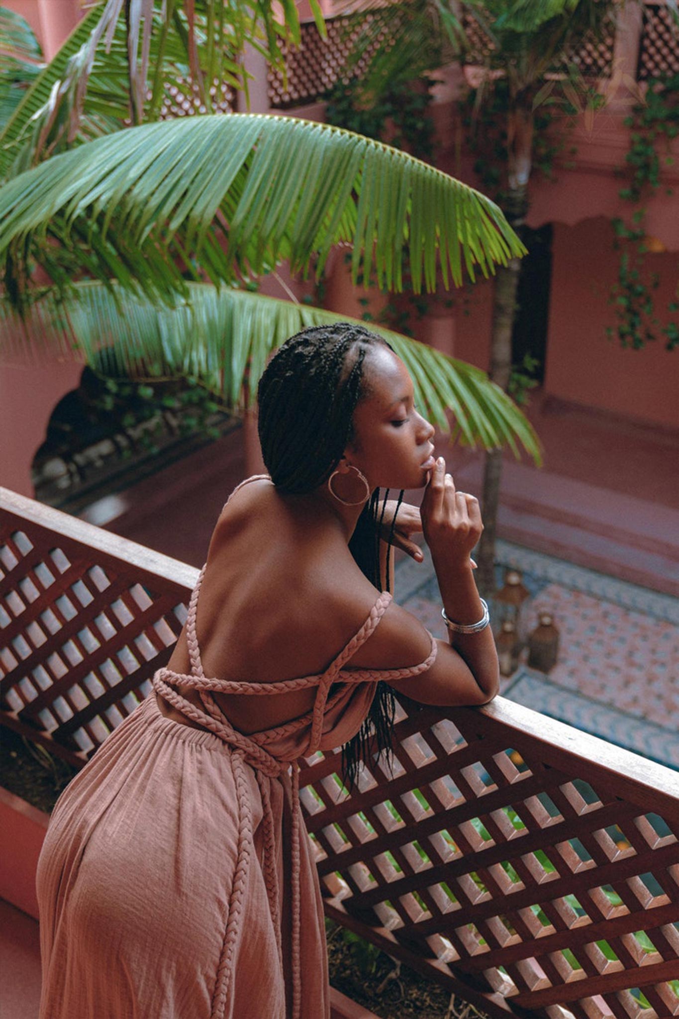 A woman with braided hair leans on a wooden railing, gazing thoughtfully into the distance. She is wearing a strappy brown dress that calls to mind the Dusty Pink Greek Goddess Prom Boho Dress by AYA Sacred Wear. The background features tropical plants and dusty pink architectural elements.