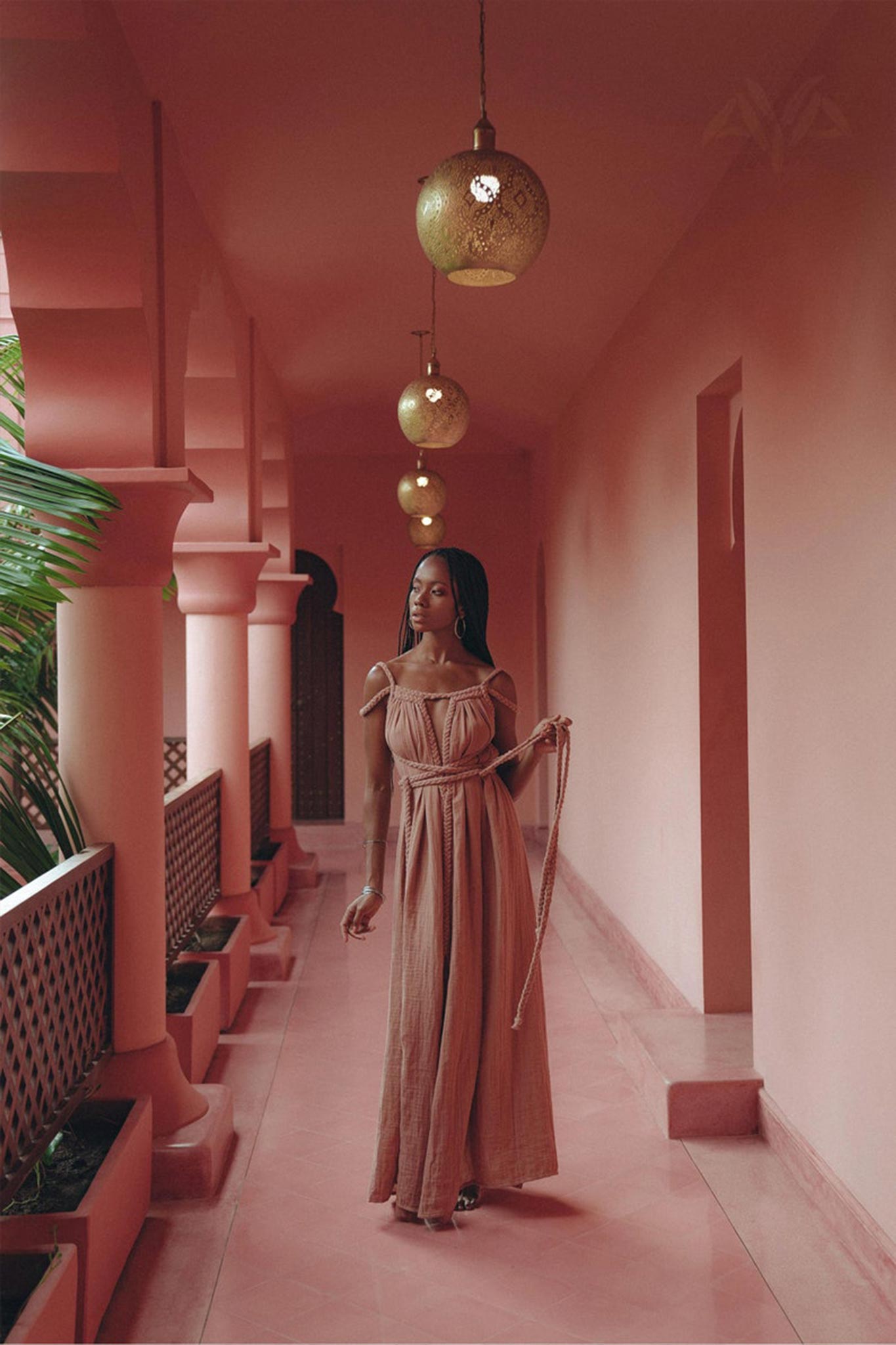 Wearing the Dusty Pink Greek Goddess Prom Boho Dress by AYA Sacred Wear, a woman gracefully strolls through a pink-hued corridor adorned with hanging golden lanterns. The walkway is framed by palm leaves and arched pillars, fostering a serene and elegant ambiance.