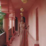 Wearing the Dusty Pink Greek Goddess Prom Boho Dress by AYA Sacred Wear, a woman gracefully strolls through a pink-hued corridor adorned with hanging golden lanterns. The walkway is framed by palm leaves and arched pillars, fostering a serene and elegant ambiance.