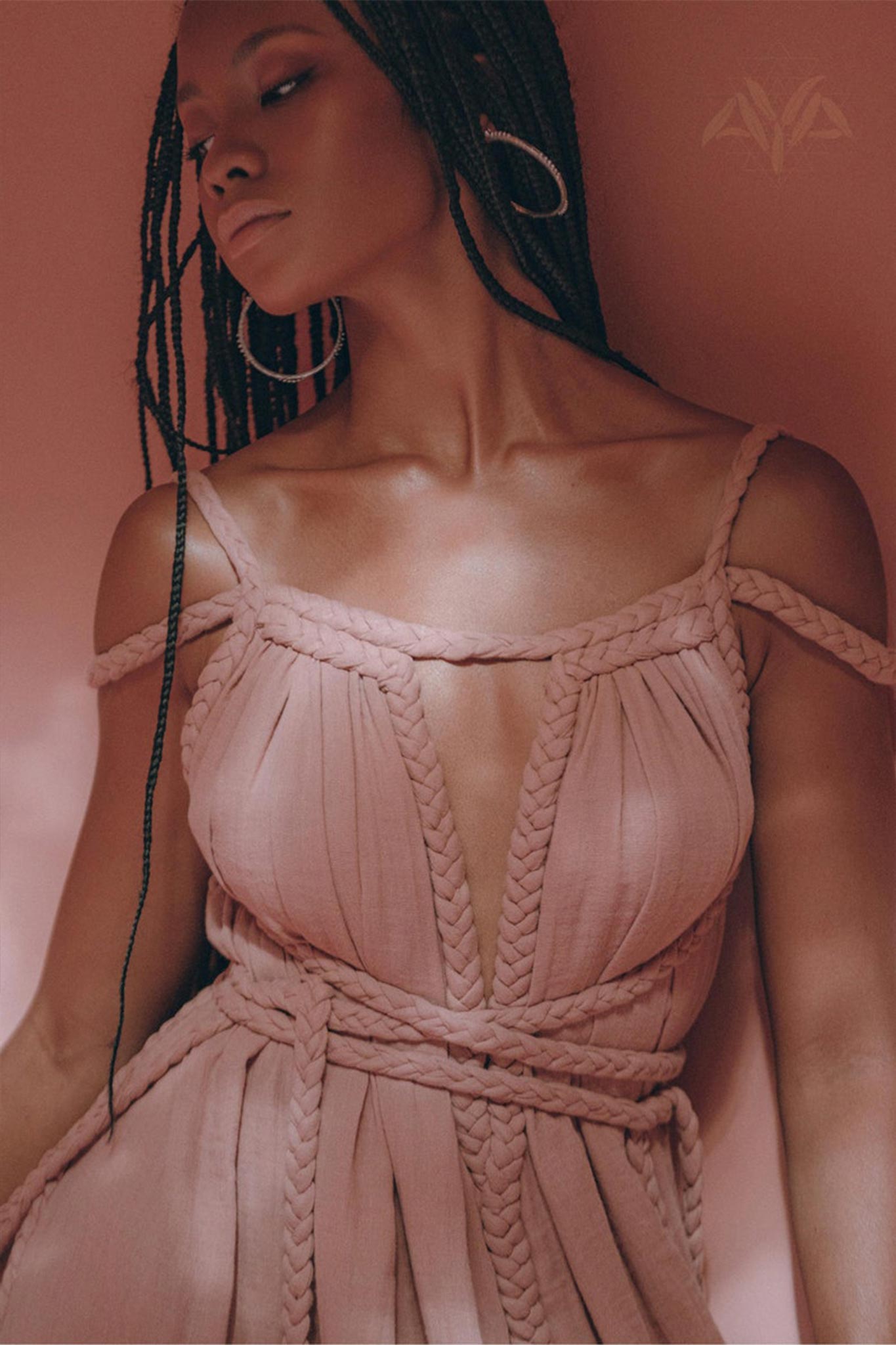 A person with long braided hair poses gracefully against a pink backdrop, showcasing an elegant and serene expression while wearing the Dusty Pink Greek Goddess Prom Boho Dress by AYA Sacred Wear.