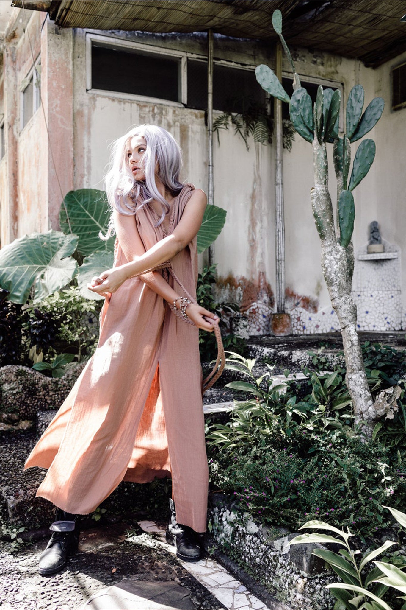 In a garden brimming with lush greenery and cacti, a person with long hair stands gracefully in a flowing Dusty Pink Nomad Spirit Dress by AYA Sacred Wear. The old building in the background lends rustic charm as they elegantly gesture with their hands.