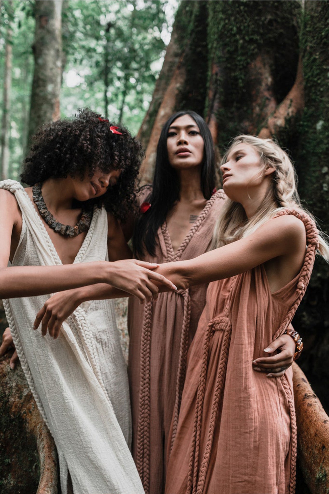 Three individuals in Dusty Pink Nomad Spirit Dresses by AYA Sacred Wear stand closely together in a lush forest setting. They exude a sense of serenity and connection, with one person gently resting their head on another's shoulder. The backdrop, filled with vibrant greenery and substantial tree trunks, further enhances the bohemian atmosphere.