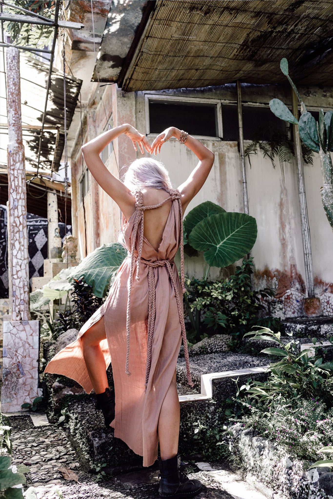 In a serene and bohemian setting, a person with white hair stands with arms raised outside a rustic, overgrown building. They are wearing the Dusty Pink Nomad Spirit Dress by AYA Sacred Wear, an adjustable size, multiway dress made of organic cotton, paired with black boots. They are surrounded by various plants and cacti.