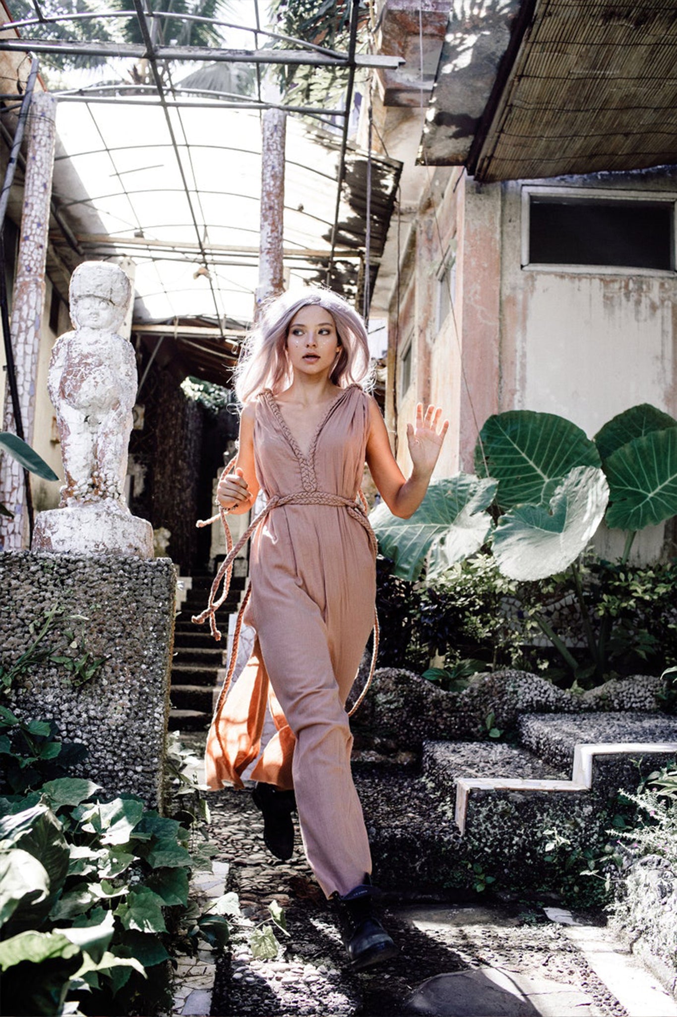 A woman wearing a Dusty Pink Nomad Spirit Dress from AYA Sacred Wear, with light-colored hair, walks energetically through a garden path surrounded by lush green leaves. She is near a weathered statue under a glass roof structure.