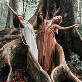 Three women in Dusty Pink Nomad Spirit Dresses by AYA Sacred Wear stand together with arms outstretched on a large tree root in a forest. Red petals are scattered on the ground, creating a serene and natural scene with the majestic tree trunk towering behind them.