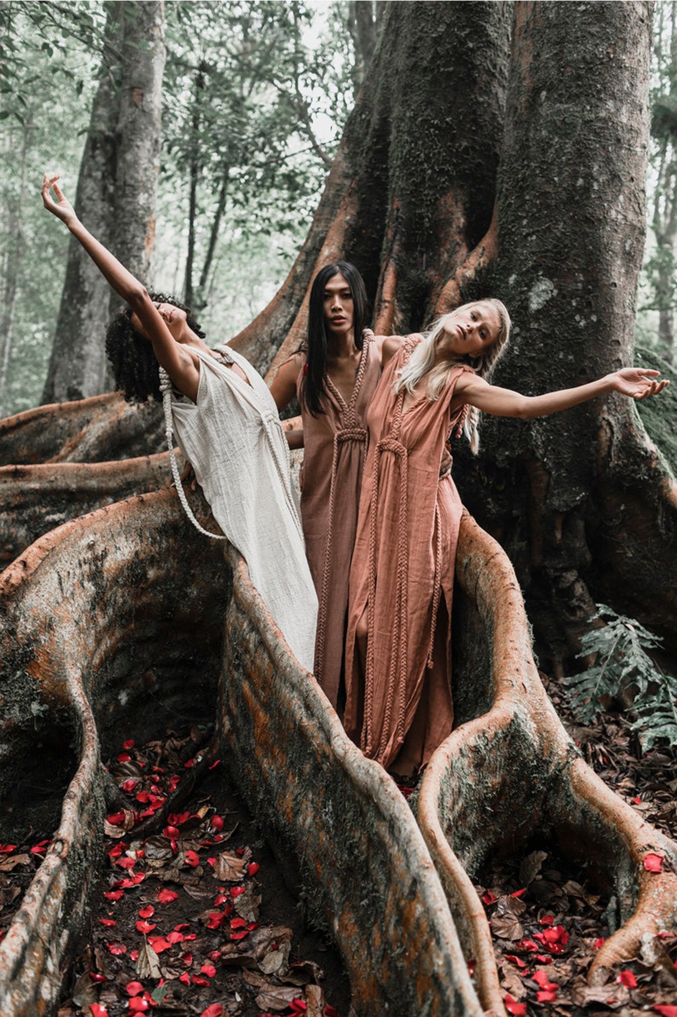 Three women in Dusty Pink Nomad Spirit Dresses by AYA Sacred Wear stand together with arms outstretched on a large tree root in a forest. Red petals are scattered on the ground, creating a serene and natural scene with the majestic tree trunk towering behind them.