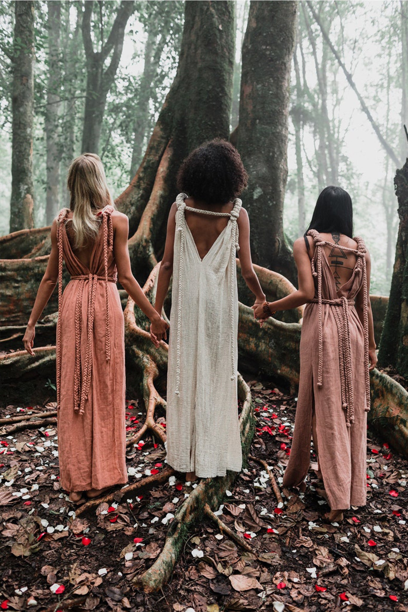 Clad in Dusty Pink Nomad Spirit Dresses from AYA Sacred Wear, three women stand hand in hand, facing away in a misty forest. Surrounded by large tree roots and scattered petals on the ground, they create a serene and mystical atmosphere.