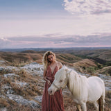 A person with long, wavy hair in an AYA Sacred Wear Dusty Rose Organic Boho Dress embellished with hand-loomed tassels stands beside a white horse on a grassy hill. The sky above is partly cloudy, and the landscape features rolling hills under a soft, pastel-colored sunset.