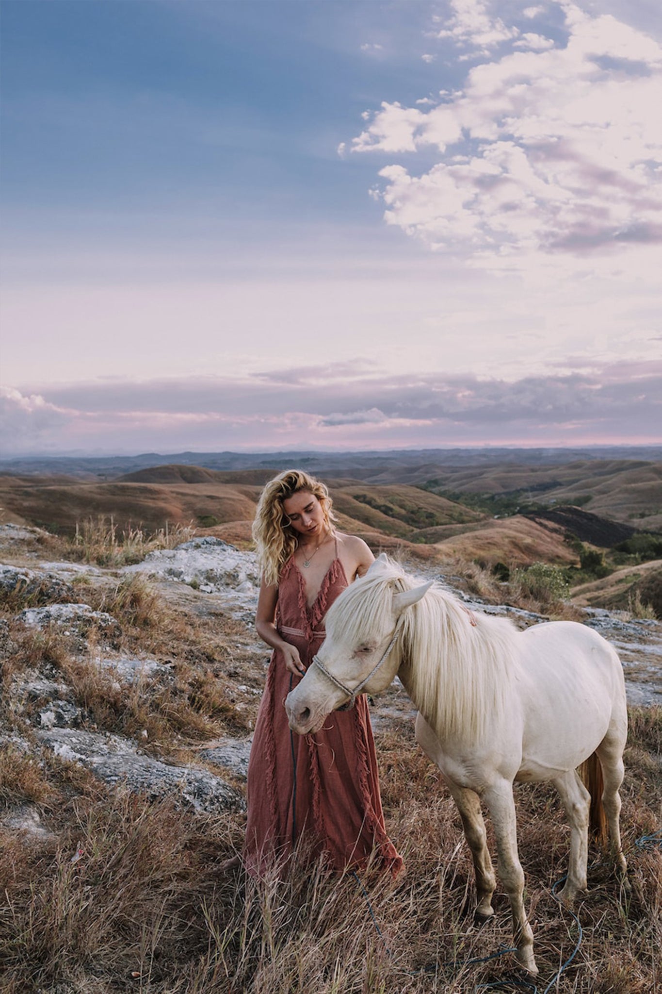 A person with long, wavy hair in an AYA Sacred Wear Dusty Rose Organic Boho Dress embellished with hand-loomed tassels stands beside a white horse on a grassy hill. The sky above is partly cloudy, and the landscape features rolling hills under a soft, pastel-colored sunset.
