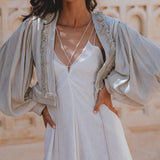 A model wearing a dusty blue puff sleeves top with organic fabric, featuring boho tassels. The top is styled over a white dress, showcasing a relaxed yet elegant look against a warm, textured background. Perfect for tropical settings.