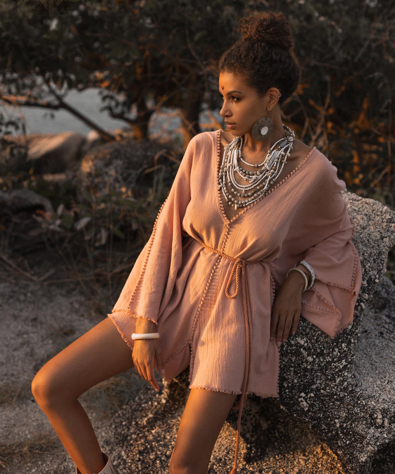 A model poses gracefully in a dusty pink handmade knotted jumpsuit romper dress, adorned with layered necklaces. The outfit features loose sleeves and a tied waist, set against a natural background of rocks and greenery, with warm light illuminating the scene, evoking a relaxed yet stylish vibe. This elegant attire is ideal for bridesmaids, capturing a goddess-like aesthetic by Aya Sacred Wear.