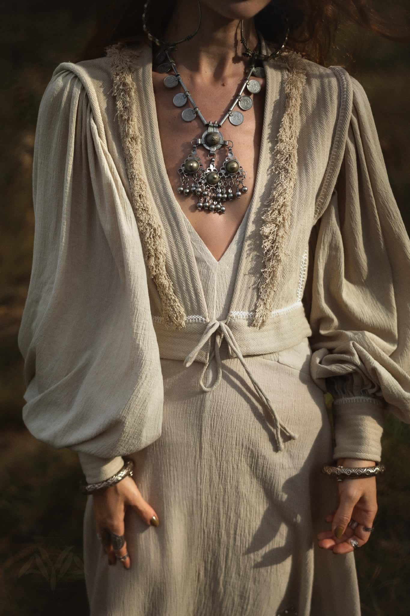 Wearing the Dusty Blue Puff Sleeves Top from AYA Sacred Wear, a person pairs this organic boho tassels crop top with an elegant jacket and large metallic statement jewelry, standing outdoors in soft lighting.