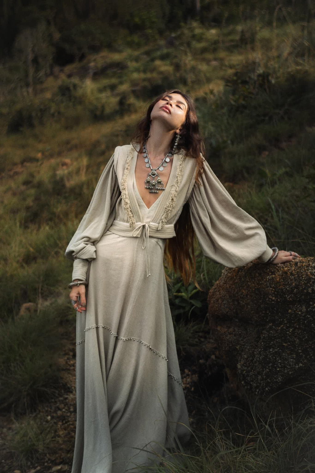 A woman stands outdoors, eyes closed, against a rock wearing the Dusty Blue Puff Sleeves Top by AYA Sacred Wear. The organic top with boho tassels highlights the grassy backdrop and distant rocks.