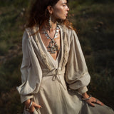 A woman stands outdoors wearing an AYA Sacred Wear Dusty Blue Puff Sleeves Top—an organic, boho tassels crop top. Her large silver necklace and earrings beautifully complement her wavy hair and hand-woven linen scarf against the lush green backdrop.