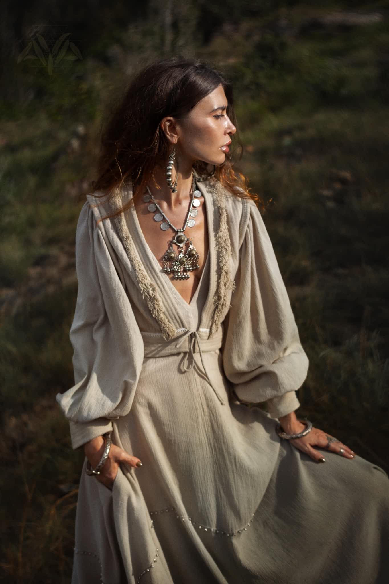 A woman stands outdoors wearing an AYA Sacred Wear Dusty Blue Puff Sleeves Top—an organic, boho tassels crop top. Her large silver necklace and earrings beautifully complement her wavy hair and hand-woven linen scarf against the lush green backdrop.