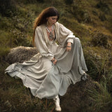 A woman in a flowing beige dress and elegant jacket sits gracefully on a rock in a grassy field. Her long hair is adorned with layered necklaces. The serene, natural ambiance perfectly complements her AYA Sacred Wear Dusty Blue Puff Sleeves Top • Boho Tassels Crop Top sophistication.