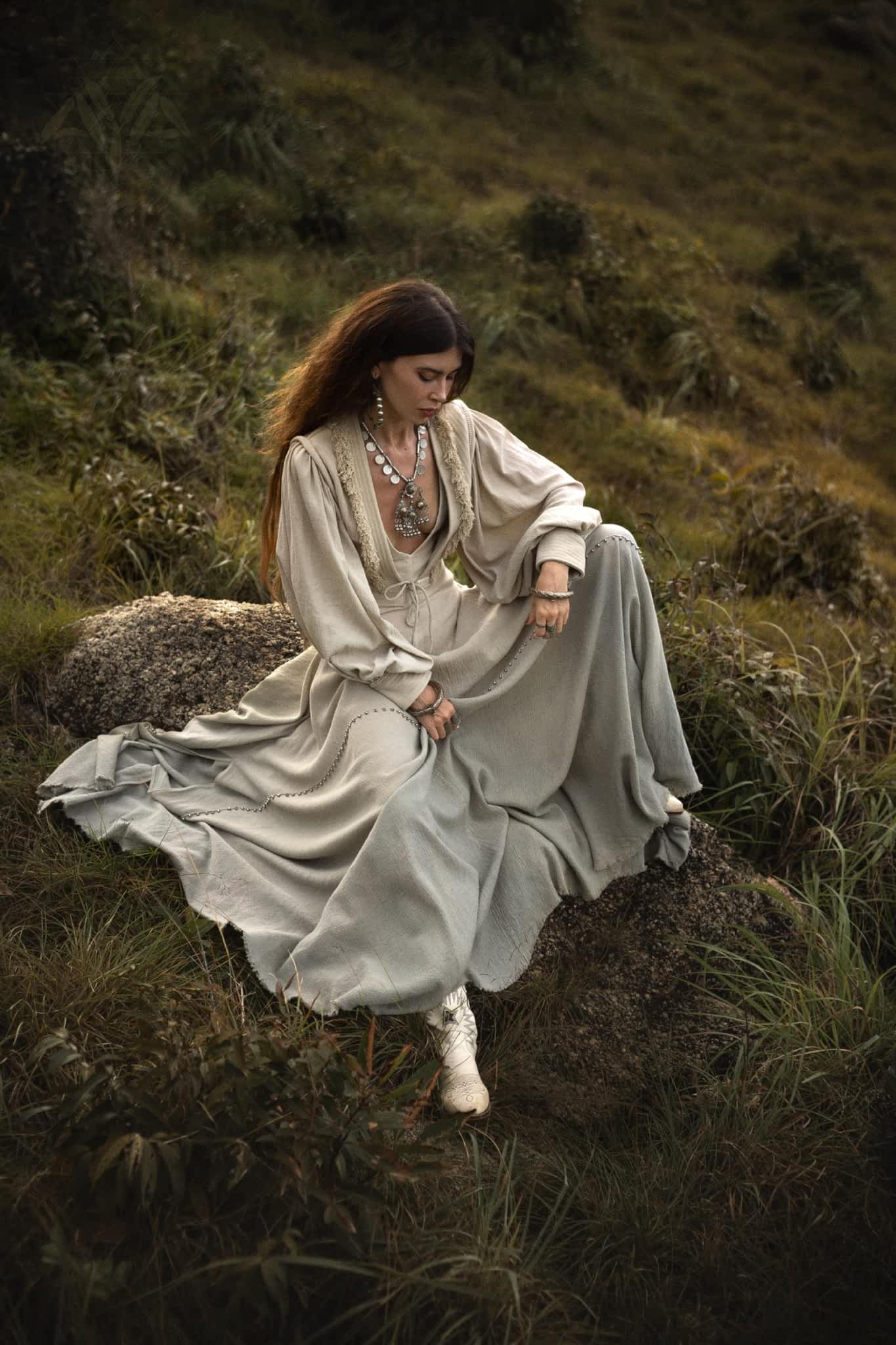 A woman in a flowing beige dress and elegant jacket sits gracefully on a rock in a grassy field. Her long hair is adorned with layered necklaces. The serene, natural ambiance perfectly complements her AYA Sacred Wear Dusty Blue Puff Sleeves Top • Boho Tassels Crop Top sophistication.