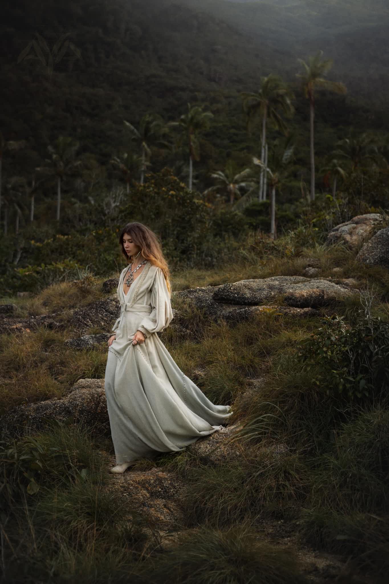 Dressed in AYA Sacred Wear's Dusty Blue Puff Sleeves Top with boho tassels, a person strolls through a lush landscape of palm trees and mountains. The scene is serene and mystical, highlighted by soft lighting and mist enhancing the background.