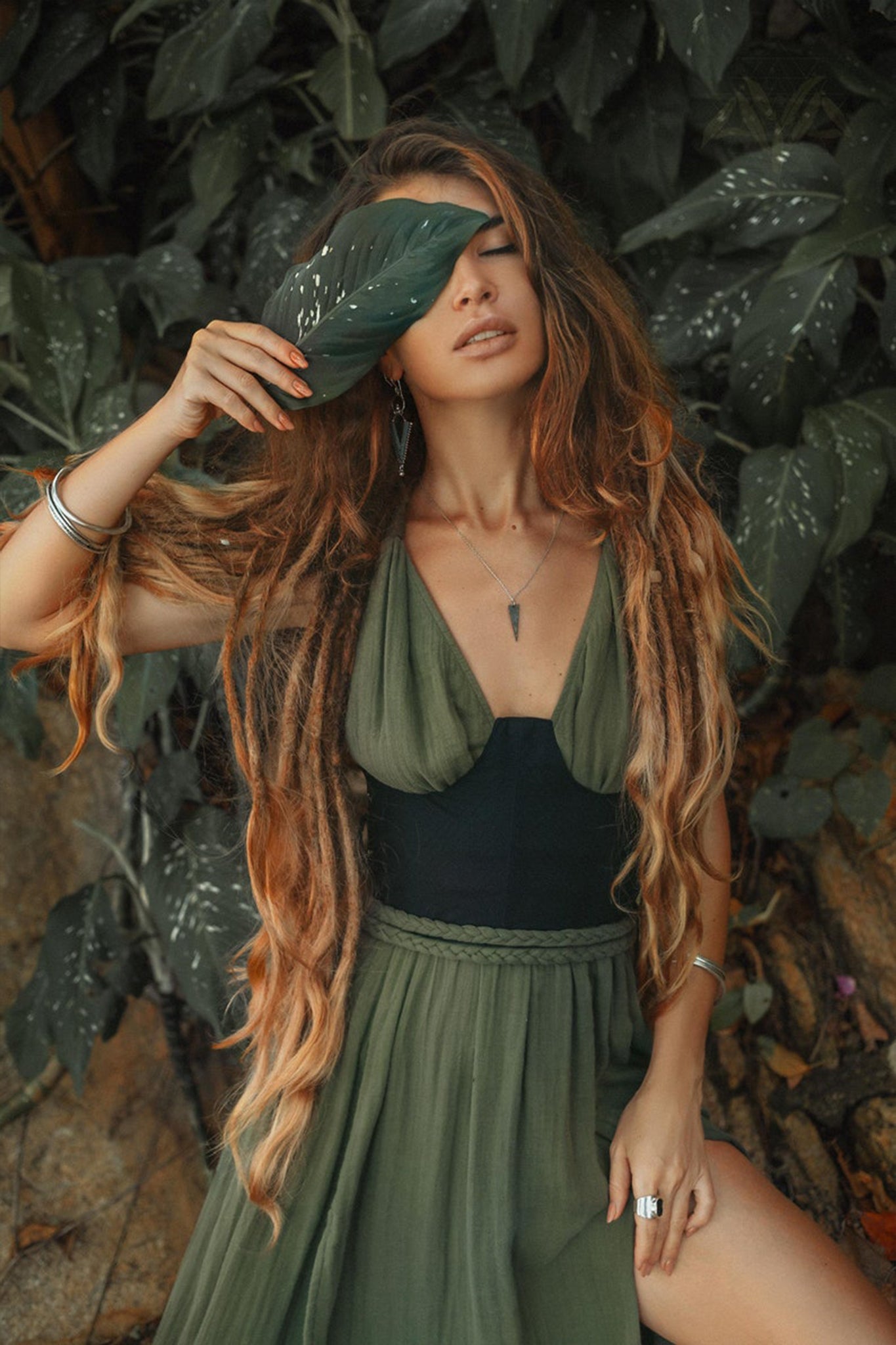 A person in an elegant, Boho Grecian Dress by AYA Sacred Wear poses gracefully, the handwoven cotton fabric enhancing their long, wavy hair. The dark green dress, botanically dyed to blend harmoniously with lush foliage, partially covers their face with a large leaf. They are adorned with silver jewelry featuring elegant rings and bracelets.