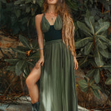A woman stands confidently on a rock, wearing the Greek Goddess Dress by AYA Sacred Wear—a boho Grecian creation crafted from handwoven cotton in dark green, featuring a high slit and paired with black boots. Surrounded by lush greenery, her wavy hair cascades down one shoulder as she gazes intently ahead.
