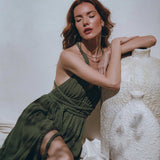 A woman in the Greek Goddess Dress by AYA Sacred Wear, a botanically dyed boho dark green masterpiece, poses with a large, textured white vase. Her long, wavy hair cascades gracefully as she sits with eyes closed, leaning on the vase. The setting is bright, with soft shadows dancing on the wall, capturing the essence of a Greek goddess.