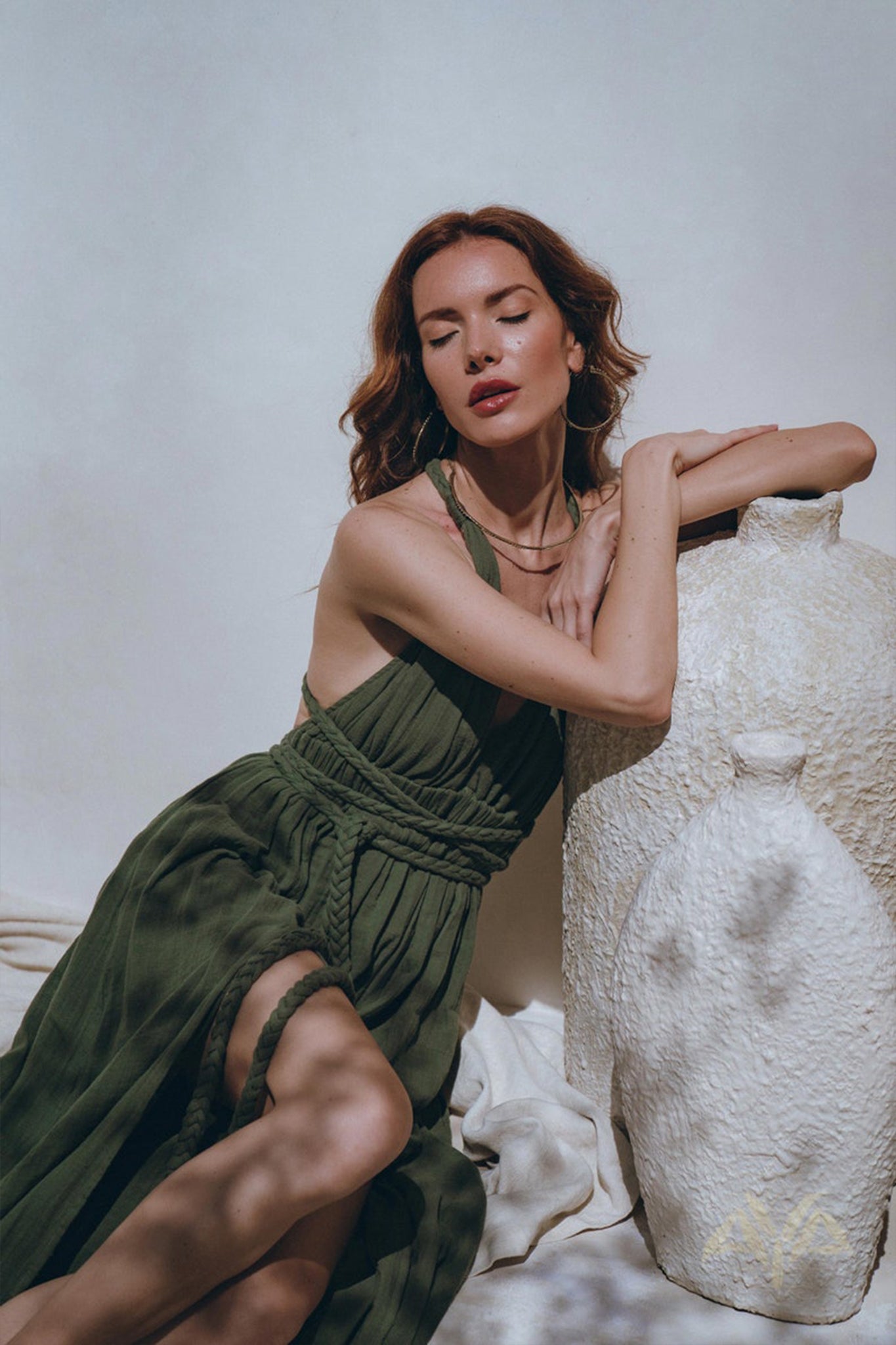 A woman in the Greek Goddess Dress by AYA Sacred Wear, a botanically dyed boho dark green masterpiece, poses with a large, textured white vase. Her long, wavy hair cascades gracefully as she sits with eyes closed, leaning on the vase. The setting is bright, with soft shadows dancing on the wall, capturing the essence of a Greek goddess.