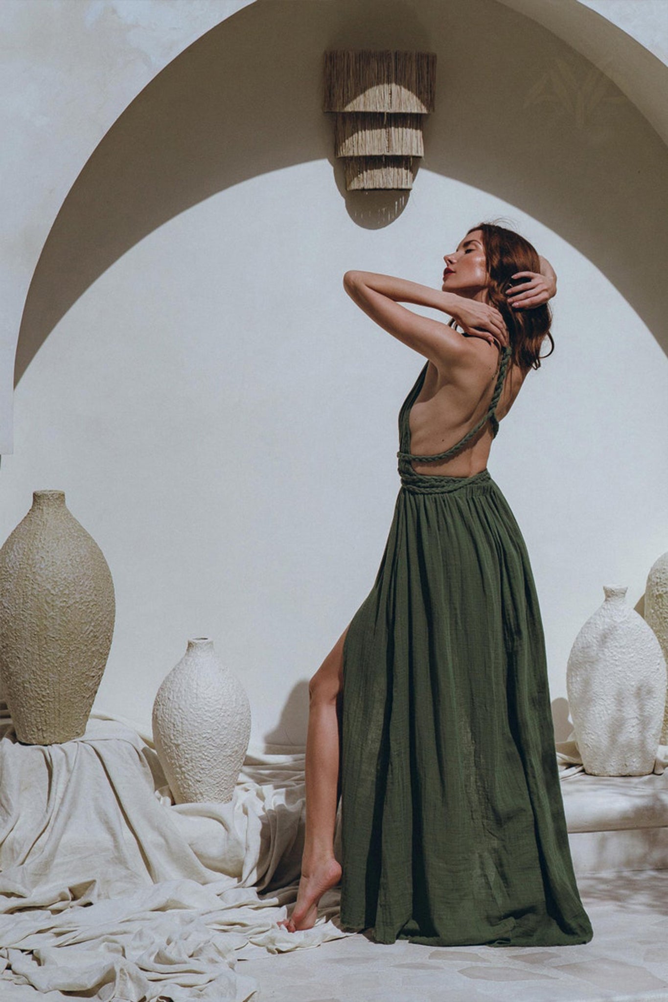 Wearing the Greek Goddess Dress by AYA Sacred Wear, a woman poses with arms raised on a textured beige surface. The scene is elegantly accented by large decorative urns and a minimalist archway, crafting a serene, artistic setting.