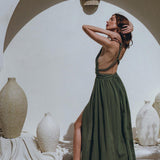 Wearing the Greek Goddess Dress by AYA Sacred Wear, a woman poses with arms raised on a textured beige surface. The scene is elegantly accented by large decorative urns and a minimalist archway, crafting a serene, artistic setting.