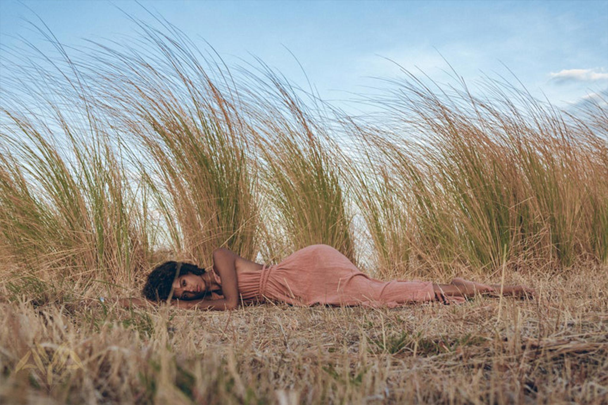 A person lies peacefully on dry grass, enveloped by tall reeds under a blue sky. They are wearing the Greek Goddess Dress in Rose Gold by AYA Sacred Wear, a flowing long maxi dress with a belt that beautifully harmonizes with the natural setting.