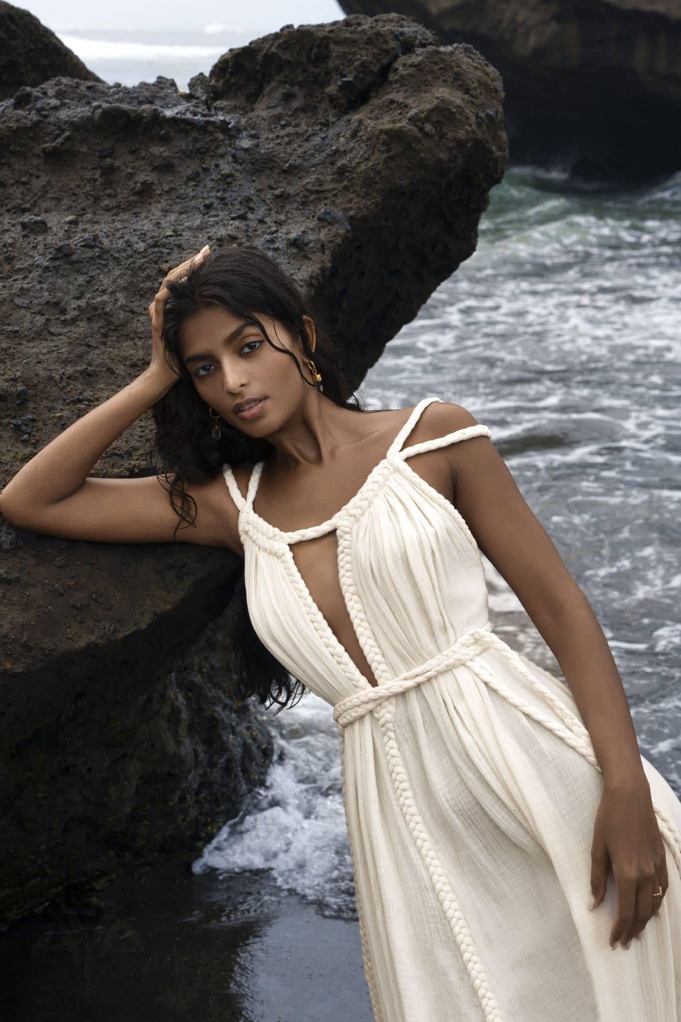 A person with long dark hair leans against a large rock by the ocean, wearing AYA Sacred Wear's Off-White Greek Goddess Boho Wedding Dress. Crafted from organic cotton with braided straps and a rope belt, it flows elegantly as sea waves crash gently in the background.