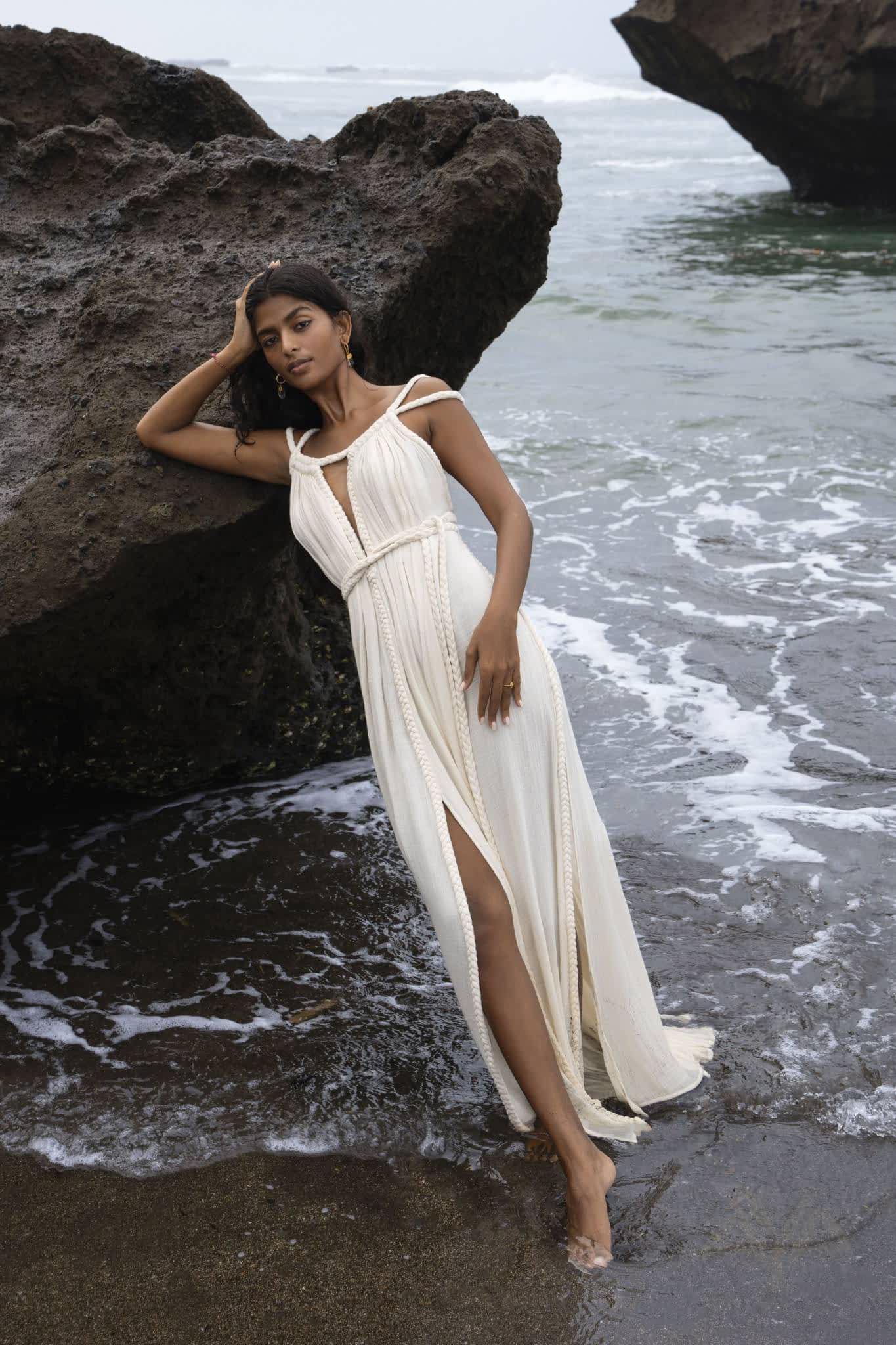 A woman wearing the Off-White Greek Goddess Boho Wedding Dress by AYA Sacred Wear leans against a large rock on the beach. The flowing dress has a slit and she stands barefoot with waves gently caressing her feet, while an overcast sky frames the serene ocean in the background.