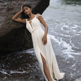 A woman wearing the Off-White Greek Goddess Boho Wedding Dress by AYA Sacred Wear leans against a large rock on the beach. The flowing dress has a slit and she stands barefoot with waves gently caressing her feet, while an overcast sky frames the serene ocean in the background.