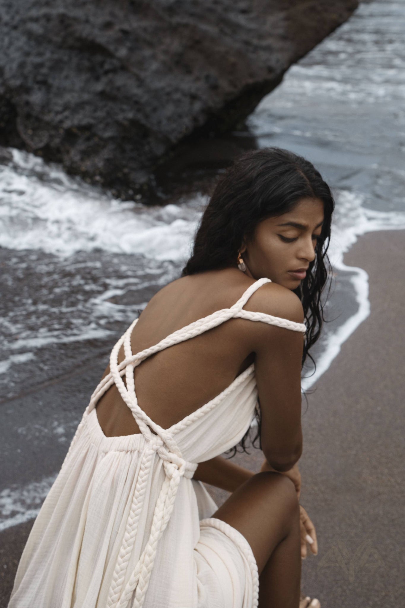 A woman with long, dark hair sits thoughtfully on a beach, clad in an Off-White Greek Goddess Boho Wedding Dress by AYA Sacred Wear. The cream-colored dress with braided straps and organic cotton complements the serene scene as waves gently wash onto the sandy shore next to a large rock.