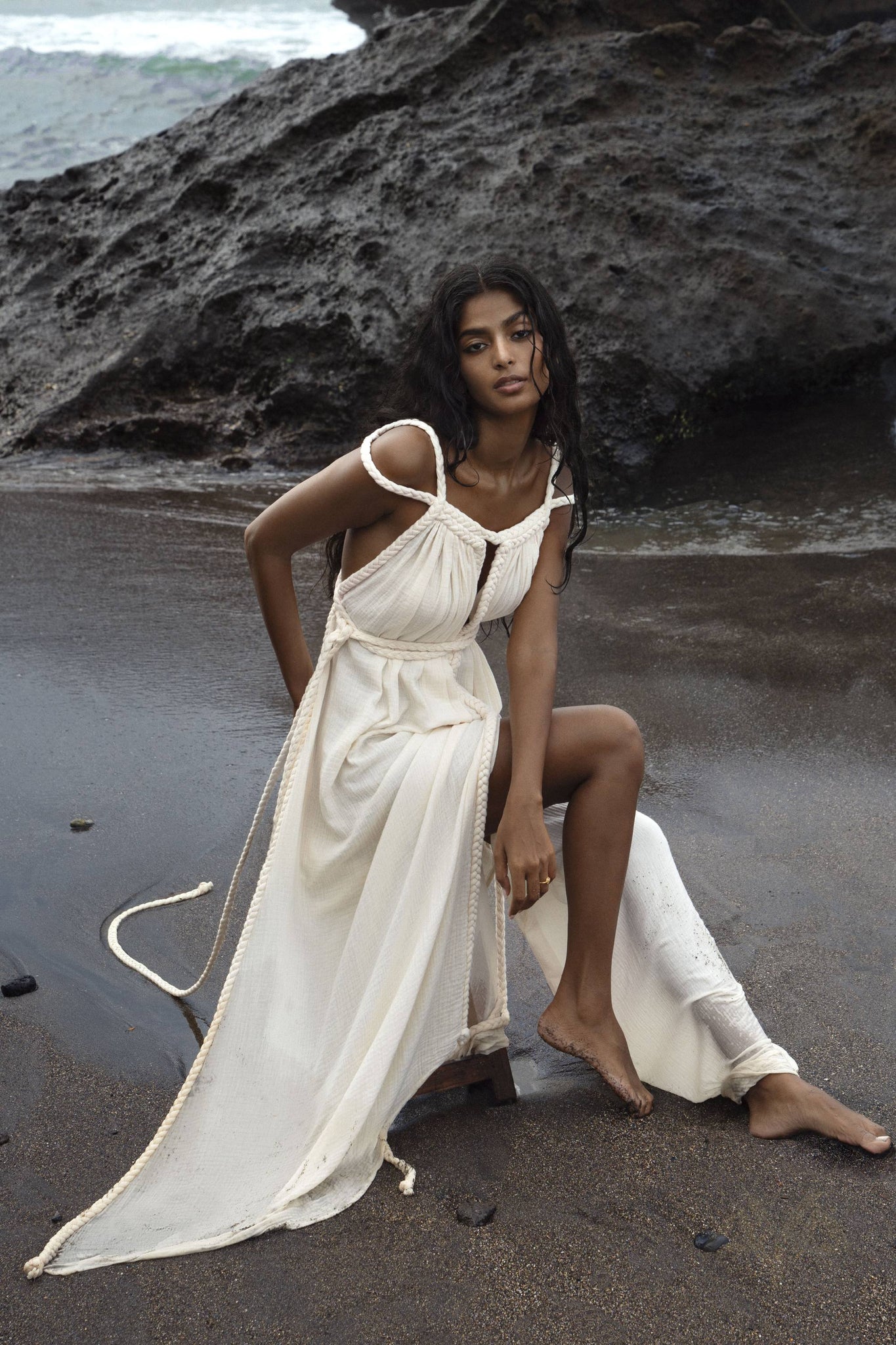 A woman with long hair sits on a rocky beach, wearing AYA Sacred Wear's Off-White Greek Goddess Boho Wedding Dress with rope-like straps. Ocean waves crash onto the dark sand as she gazes at the camera, exuding serene beauty and handmade elegance.