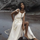 A woman in an Off-White Greek Goddess Boho Wedding Dress by AYA Sacred Wear sits on a rocky beach. Her long hair falls freely as she gazes at the camera, with the ocean behind her and dark, wet sand beneath, highlighting the organic cotton beauty of her boho wedding dress.