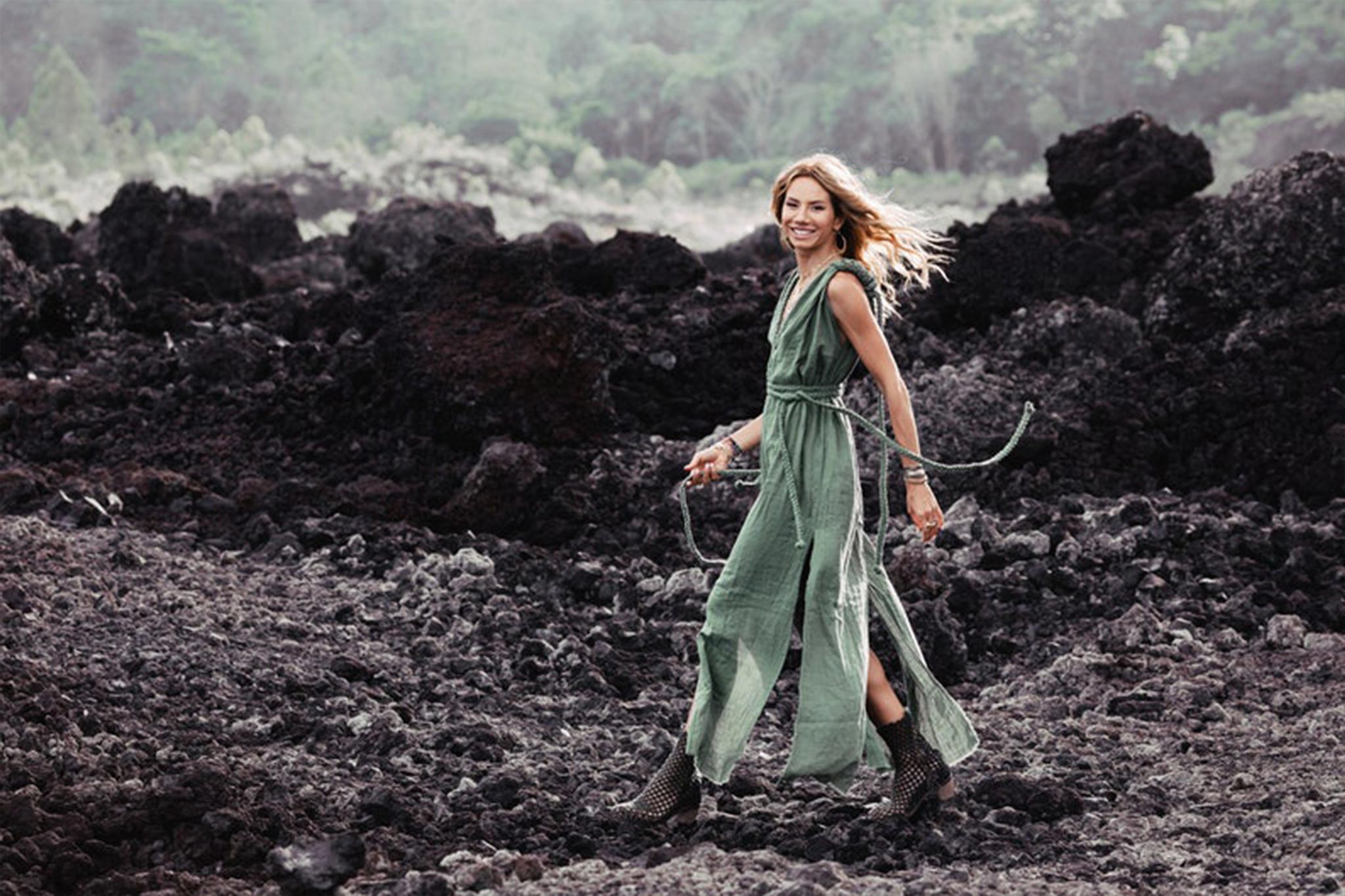 A woman in an Aya Sacred Wear Green Nomad Spirit Dress strolls joyfully over rocky terrain, surrounded by dark volcanic rocks and lush greenery in the background. She wears boots, and her hair flows in the breeze.