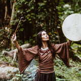 In a forest, a woman holds a drum and mallet, dressed in a brown top with flowing sleeves and a necklace. She wears the Grey Cotton Boho Skirt from AYA Sacred Wear, made of organic cotton. Lush green foliage surrounds her as she gazes upward with focus.