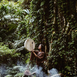 Standing on a fallen tree in a dense, lush forest, a person plays a large drum while clad in AYA Sacred Wear's Grey Cotton Boho Skirt. Crafted from organic cotton, the flowing fabric complements the smoke that gently rises around them, creating a mystical atmosphere amidst the greenery and trailing vines. The scene evokes a deep sense of harmony with nature.