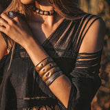 A woman is standing outdoors wearing a Hand Loomed Cotton Blouse by AYA Sacred Wear. She has long hair and complements her look with a chunky bead necklace, wide metal bangles, and a ring. The warm lighting accentuates the earthy tones of the scene, while her sustainable bohemian top enhances the natural ambiance around her.