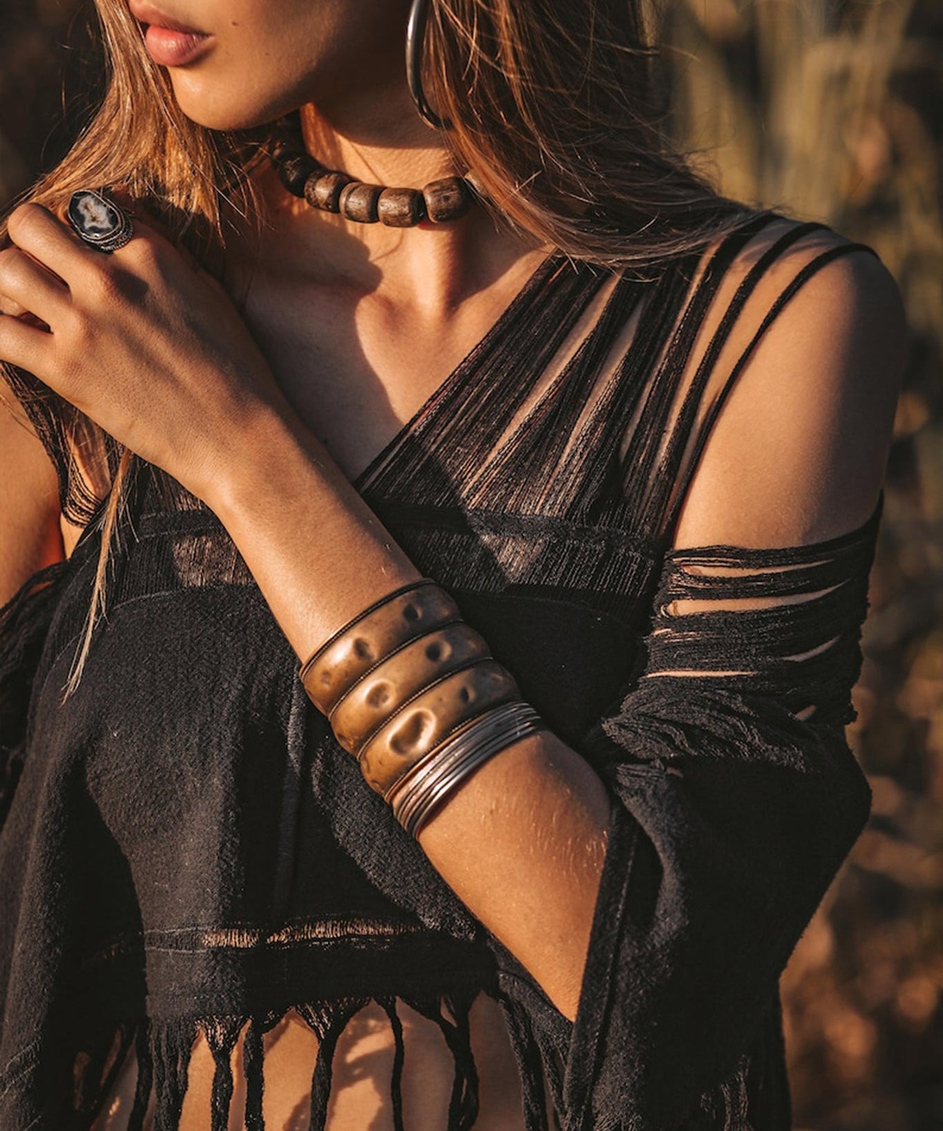 A woman is standing outdoors wearing a Hand Loomed Cotton Blouse by AYA Sacred Wear. She has long hair and complements her look with a chunky bead necklace, wide metal bangles, and a ring. The warm lighting accentuates the earthy tones of the scene, while her sustainable bohemian top enhances the natural ambiance around her.