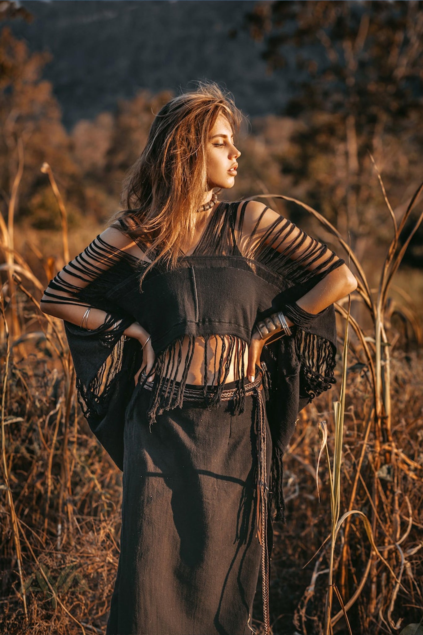 A woman stands confidently in a field, wearing the Hand Loomed Cotton Blouse by AYA Sacred Wear, a black bohemian top with fringes. Her arms are raised, and her hair flows gracefully as the warm sunlight casts a golden hue over the tall grasses and blurred trees in the background.