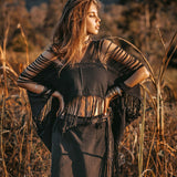 A woman stands confidently in a field, wearing the Hand Loomed Cotton Blouse by AYA Sacred Wear, a black bohemian top with fringes. Her arms are raised, and her hair flows gracefully as the warm sunlight casts a golden hue over the tall grasses and blurred trees in the background.