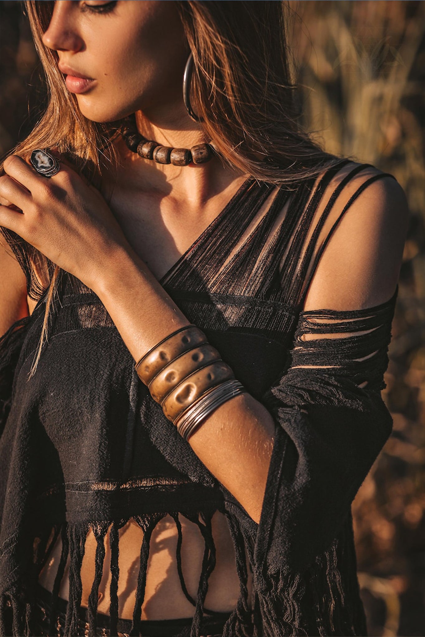 A woman is standing outdoors wearing a Hand Loomed Cotton Blouse by AYA Sacred Wear. She has long hair and complements her look with a chunky bead necklace, wide metal bangles, and a ring. The warm lighting accentuates the earthy tones of the scene, while her sustainable bohemian top enhances the natural ambiance around her.