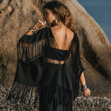 A woman wearing the AYA Sacred Wear Hand Loomed Cotton Blouse stands on a pebbly beach, facing a large rock. Her back is exposed, and she has long, wavy hair. The sky is clear and blue, reflecting the serenity of her surroundings.