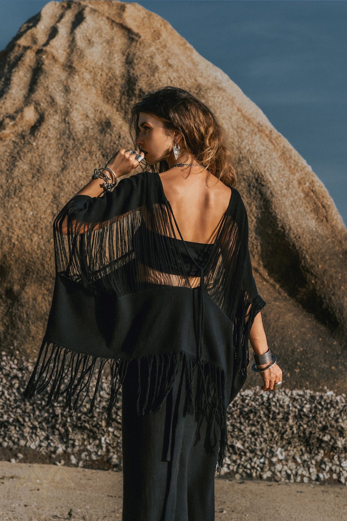 A woman wearing the AYA Sacred Wear Hand Loomed Cotton Blouse stands on a pebbly beach, facing a large rock. Her back is exposed, and she has long, wavy hair. The sky is clear and blue, reflecting the serenity of her surroundings.