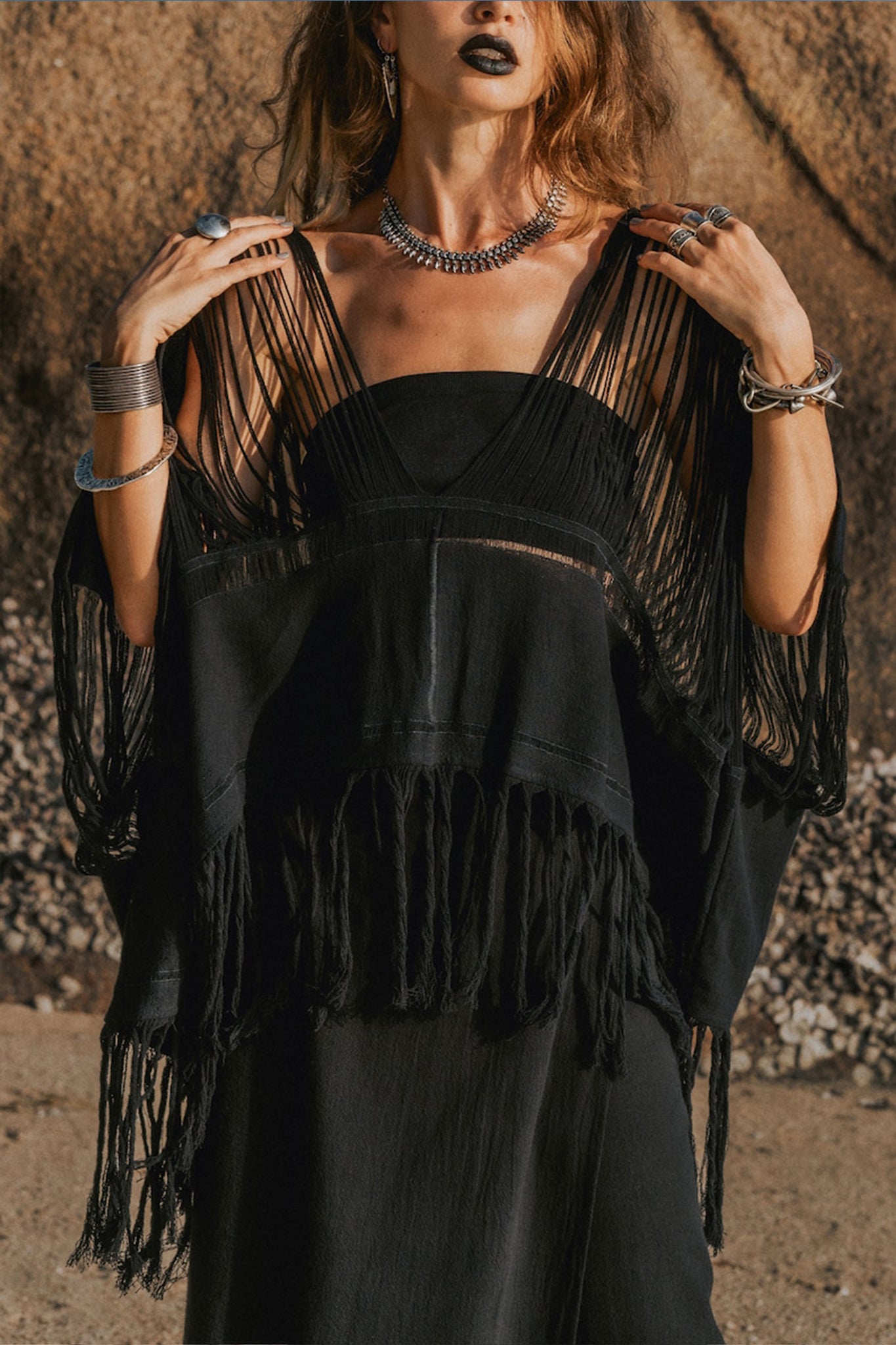 A person wearing a dark, fringed poncho over an AYA Sacred Wear Hand Loomed Cotton Blouse, beautifully paired with bold jewelry like rings, bracelets, and a necklace. They stand outdoors against a rugged rock background, with tousled hair and dark lipstick.